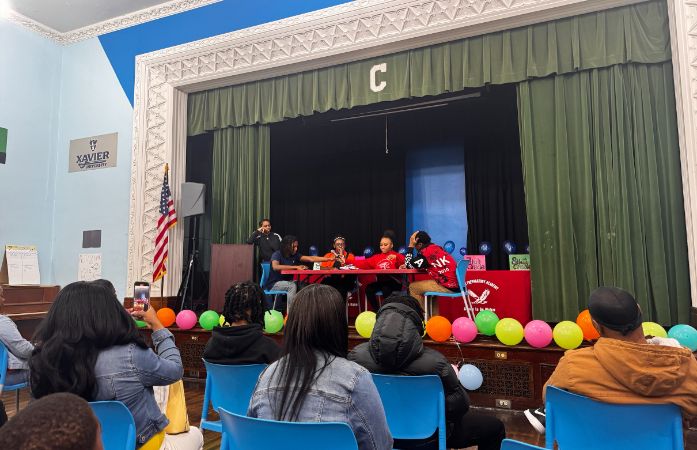  Eighth grade students sit at a table on stage performing a Black history skit for the audience duri